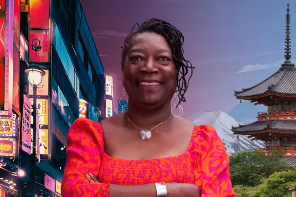 Woman with brown braids wearing red jumpsuit with arms crossed smiles at camera with scene of Japan behind her.