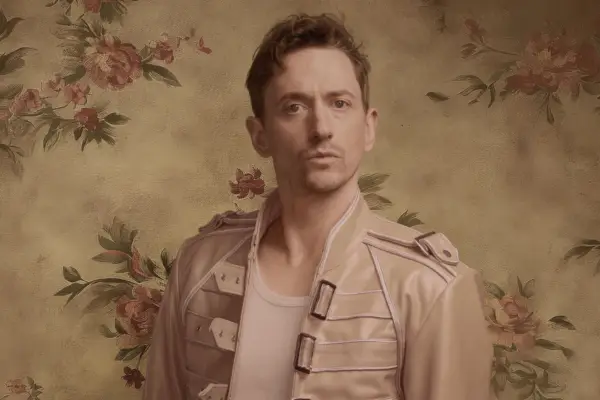 John Robins in a white t-shirt and cream jacket looking ahead. He has short hair and light stubble.