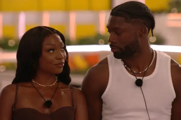 Mimii and Josh looking at each other, both smiling. Mimii is wearing a brown dress with mic around her neck while Josh is wearing a white vest and also has a mic around his neck.