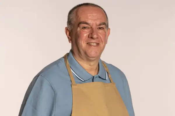 Bake Off 2023 contestant Keith wearing a blue polo and brown apron, smiling into camera