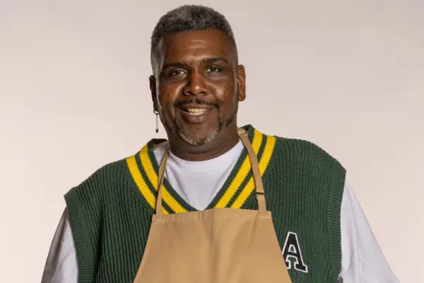 Bake Off 2023 contestant Amos wearing a green and yellow sleeveless sweater and brown apron, smiling into camera