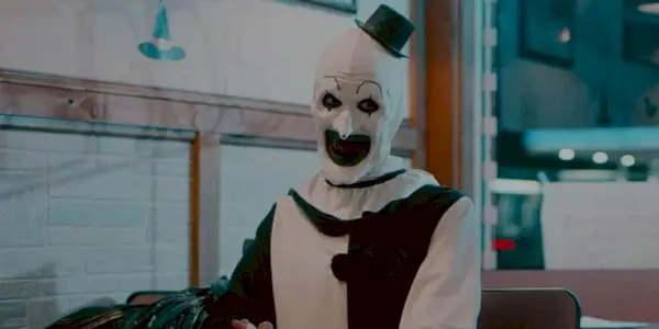 David Howard Thornton as Art the Clown sitting in a pizza parlor smiling oddly in Terrifier