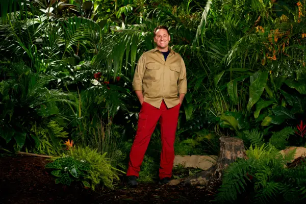 Nick Pickard dressed in a khaki shirt and red trousers while posing in front of some trees for I