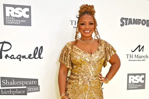 Marisha Wallace in a gold dress with one hand on her hip. She is smiling in front of a white background with words on