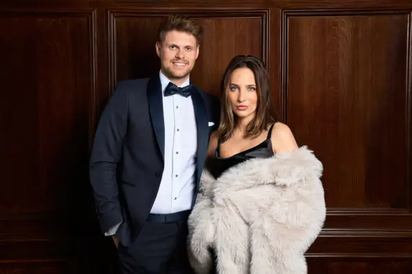 James and Maeva posing for a photo for Made in Chelsea. James is in a tuxedo and has his hands in his pockets as he smiles while Maeva is wearing a black dress and fur coat.
