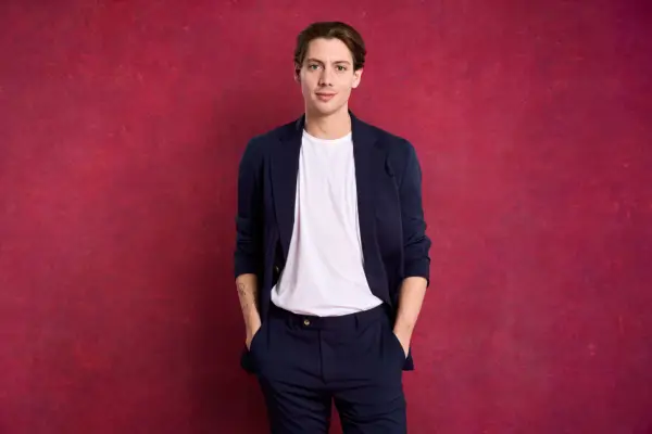 A man wearing a blue suit and white t-shirt, looking ahead with his hands in his pockets.
