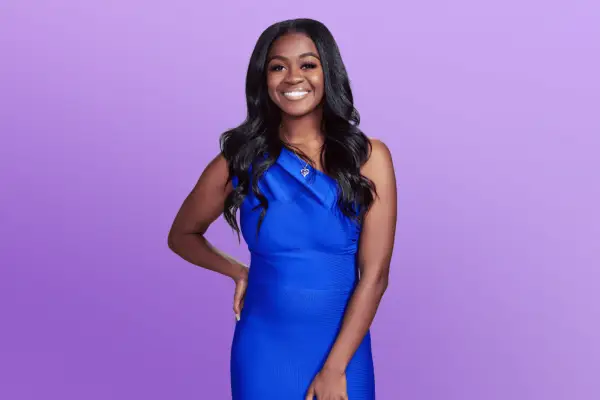 Danielle in along blue dress posing in front of a purple background with her hand on her hip