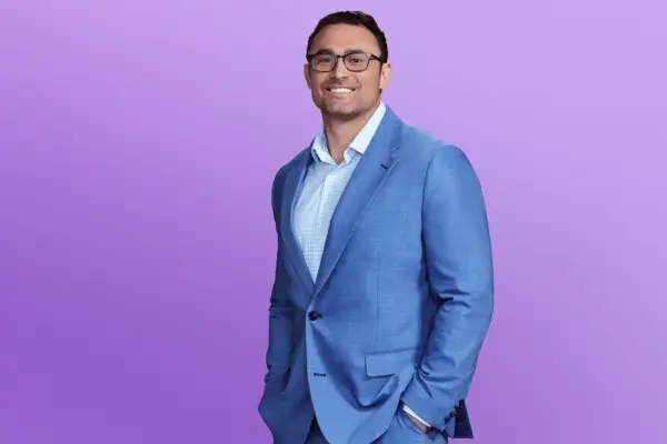 Matthew is in an all-blue suit posing in front of a purple background with his hands in his pocket