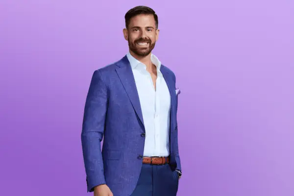 Vince in a blue and white suit with one hand in his pocket. He is posing in front of a purple background