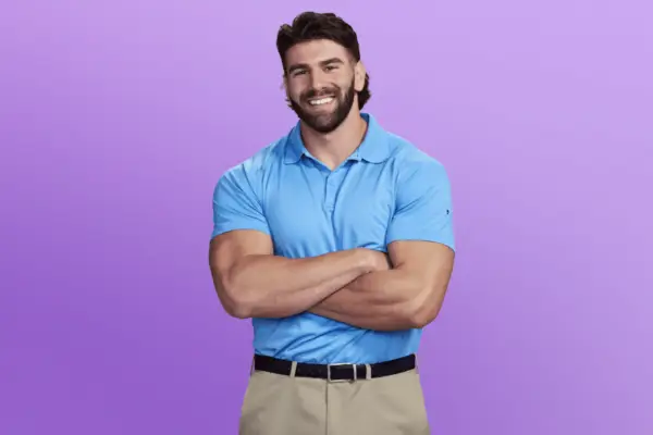 Trevor in a blue polo shirt and khaki trouser. He has his arms folded and is posing in front of a purple background