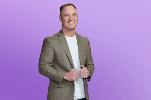 Jimmy in a green blazer and blue jeans holding onto the lapels. He is posing in front of a purple background