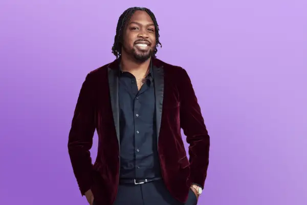 Kenneth is in a red and black suit posing in front of a purple background with his hands in his pocket