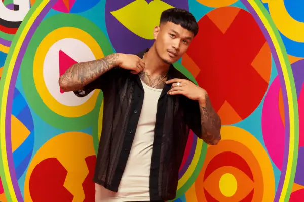 Zak holding the collar of his shirt and standing in front of the Big Brother eye