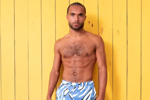 Reuben stood in front of a yellow background, with his arms by his sides looking ahead. He is topless and is wearing blue and white swim shorts.