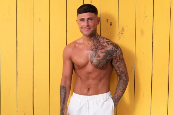 Harry stood in front of a yellow wooden background, wearing white swimshorts with one hand in his pocket. He has tatoos across his left arm and chest.