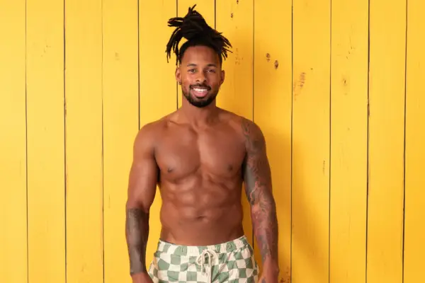 Konnor Ewudzi posing in front of a yellow wooden background. He is topless and is wearing white and green checkered shorts.