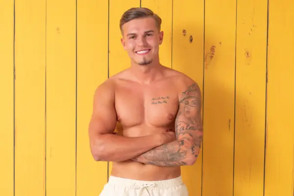 Hugo smiling as he poses in front of a yellow wooden background, topless with his arms folded in front of him.