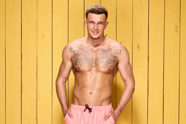 Wil Anderson posing in front of a yellow wooden panel for Love Island. He is topless and is wearing pink swim shorts. His hands are in his pockets and he is smiling.