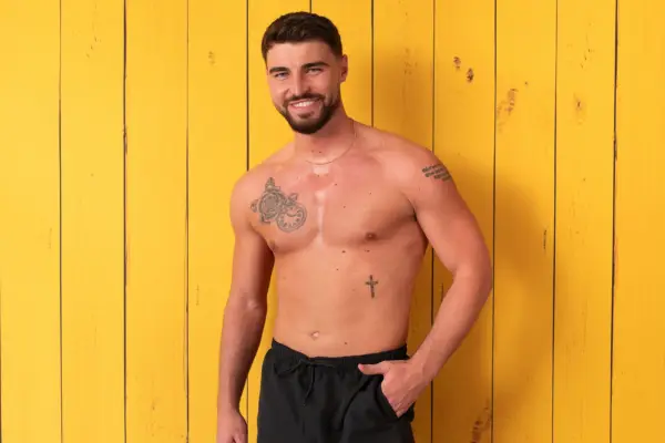 Trey stood in front of a yellow wooden background, smiling ahead. He is topless and is wearing black swim shorts and has his hand in his pocket.