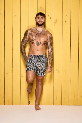 Sam Taylor poses in swim shorts in front of a yellow wooden wall for Love Island 2024.