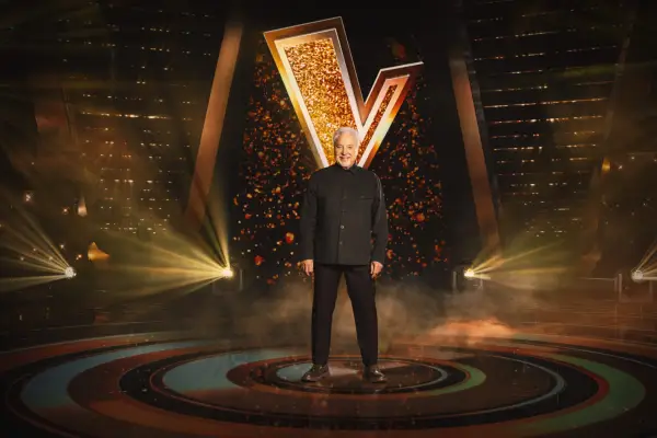 Sir Tom Jones stood in front of a golden The Voice logo, wearing an all-black suit.