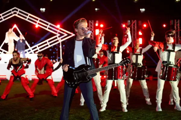 Tom Fletcher wearing a white t-shirt, black blazer and jeans and holding a microphone and guitar.