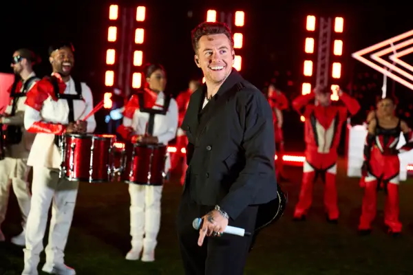 Danny Jones wearing a black shirt, smiling and holding a microphone with a band performing behind him.
