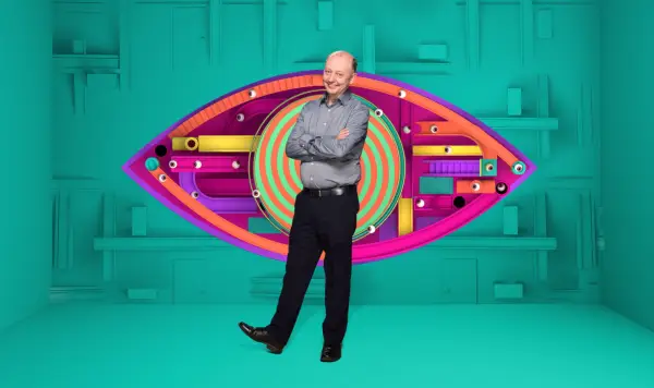 Richard wearing a grey shirt and black trousers, smiling ahead with his arms folded.