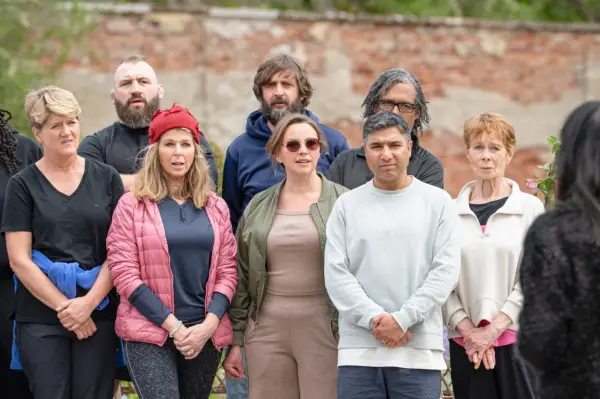 The Cast of The Celebrity Traitors stand as a group listening to host Claudia Winkleman explain the rules of a mission.