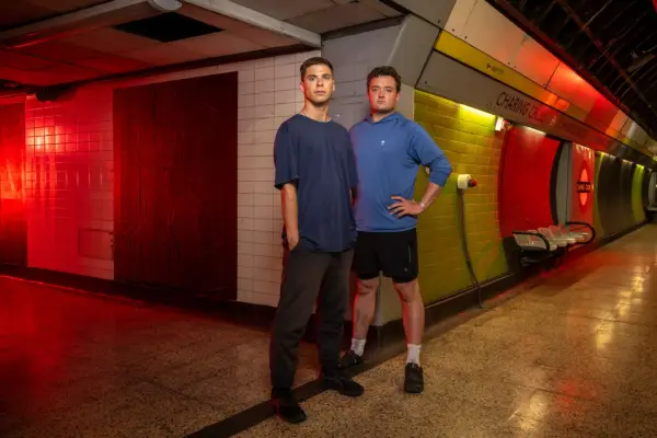 Alex and Jack stood in exercise clothing in a dimly lit Charing Cross station. They are stood next to each other while looking ahead