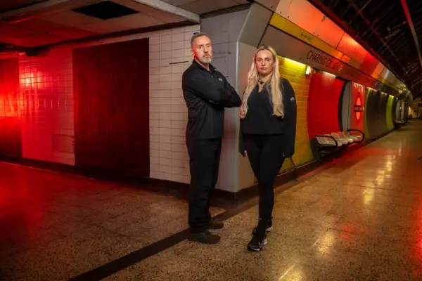 Steve and Beth stood in exercise clothing in a dimly lit Charing Cross station. They are stood next to each other while looking ahead
