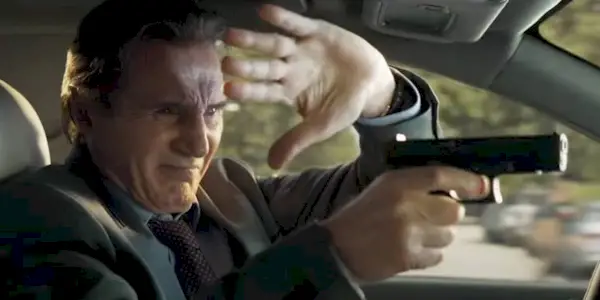 Frank Drebin Jr. (Liam Neeson) blocking his eyes from sunlight while aiming his gun, while driving, in The Naked Gun.