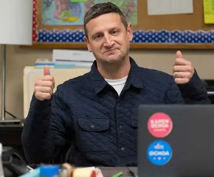 Tim Robinson looking forward with two thumbs up and sitting at a desk in I Think You Should Leave