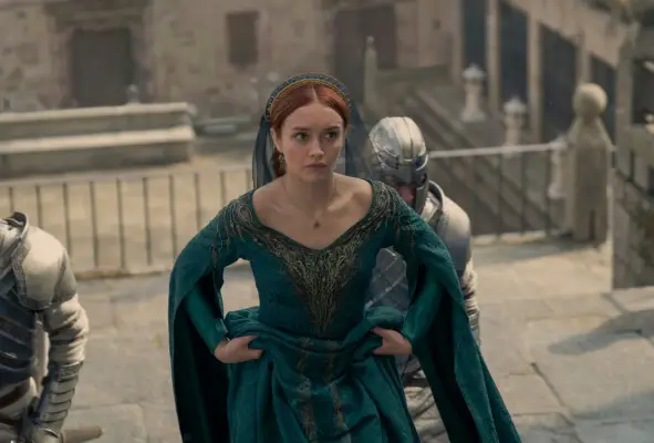 Olivia Cooke as Dowager Queen Alicent Hightower walks up stairs holding her green dress and wearing a green veil with a knight walking behind her in House of the Dragon