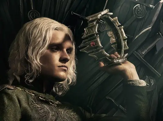 Tom Glynn-Carney as King Aegon II Targaryen sitting atop the Iron Throne holding a crown in his hand for House of the Dragon season 2.