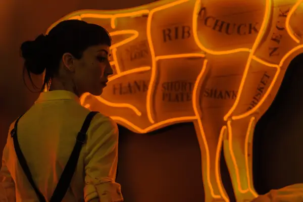 Briana Cuoco as Jenny Green in Dead Boy Detectives, stood next to a neon wall sign of a cow