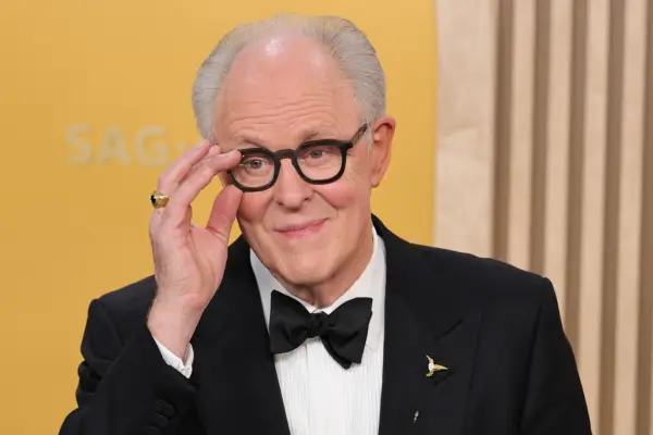 John Lithgow wearing a tuxedo, smiling ahead, as he holds his glasses.