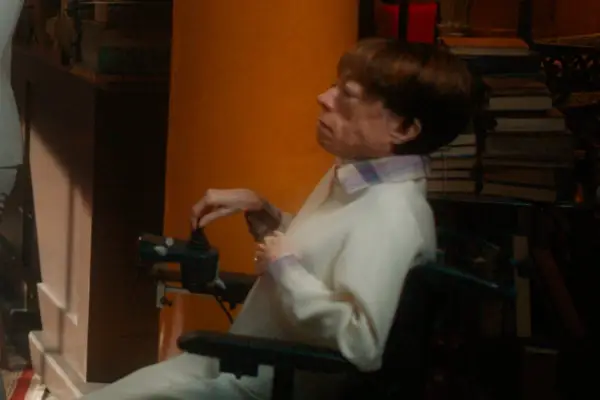 Liz Carr in Good Omens season 2, wearing a chequered shirt and white jumper