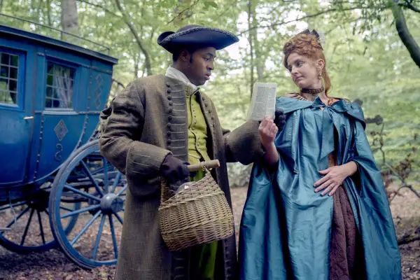 Ényì Okoronkwo as Rasselas and Louisa Harland as Nell Jackson in Renegade Nell. They are stood by a carriage, wearing period dress, and she is showing him a piece of paper