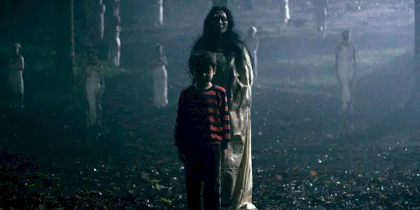 A young boy and a woman in white stand in the meadow with silhouettes of other ghostly women in white visible behind them in Satan