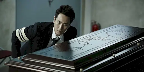 Lee Sun-kyun wearing a suit and a black and white mourning ribbon on his right arm, leaning toward a closed casket with his left ear