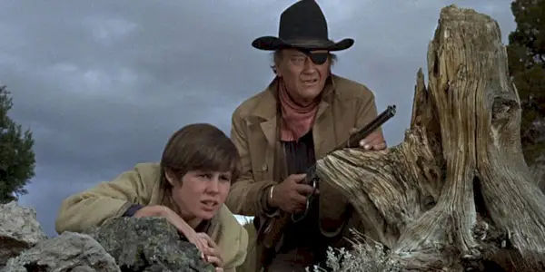 Kim Darby and John Wayne with a shotgun crouching down behind rocks looking out at something inTrue Grit