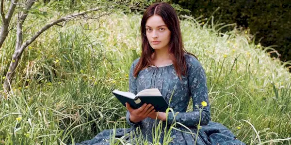 Emma Mackey as Emily reading in the field in Emily (2023)