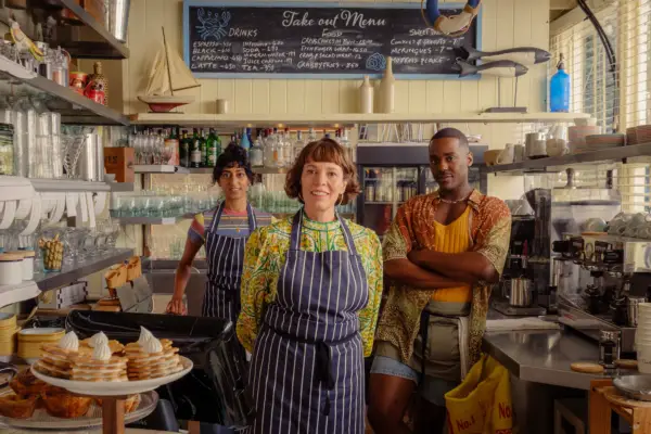 Sunita Mani, Olivia Colman, and Ncuti Gatwa in The Roses standing in a café and smiling.