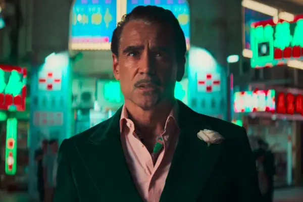 Colin Farrell in Ballad of a Small Player, wearing a dark velvet suit with a pale pink shirt and white boutonniere, standing against a backdrop of glowing neon signs in a bustling Asian night market street.