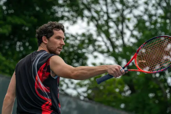 Josh O’Connor stars as Patrick wesring a red and black vest and holding a matching tennis racket in director Luca Guadagnino’s CHALLENGERS