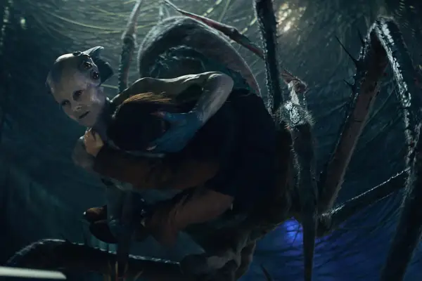 Harmada – a giant spider-like alien with a humanoid head and torso – descends from above, carrying someone in their arms