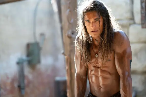 Tarak stands up straight, leaning on a table or work surface, with his shirt off