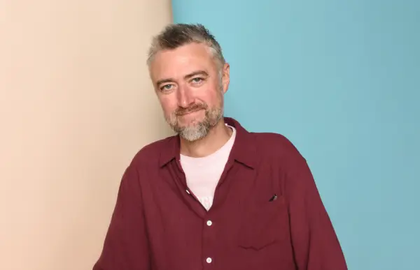 Sean Gunn smiles for the camera in a studio setting, with a beige and blue backdrop behind him