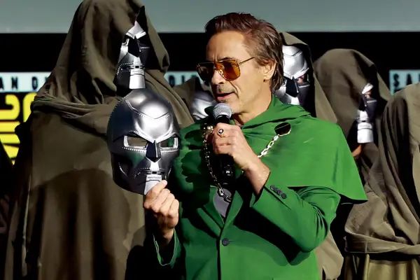 Robert Downey Jr holding a Doctor Doom mask and wearing green robes and sunglasses on stage at Comic-Con. He is talking into a microphone.